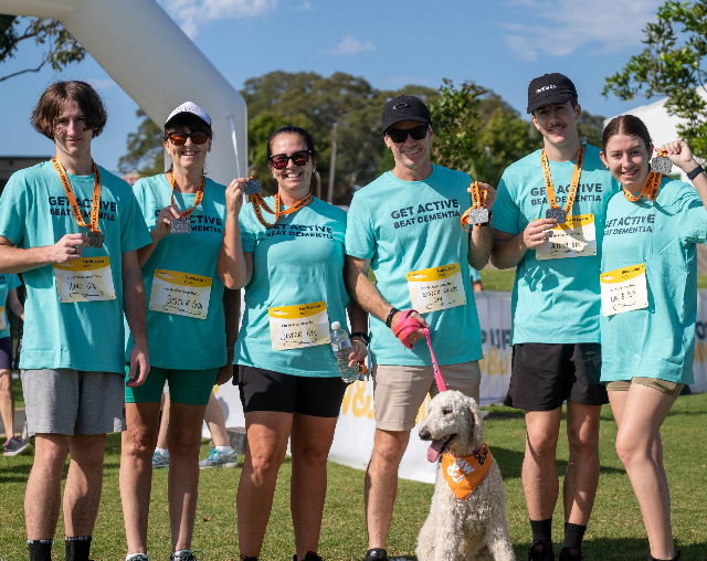 Memory Walk & Jog. Family of six people with one wearing teal shirts standing at finish line holding medals. One family member is holding a white dog on a leash.